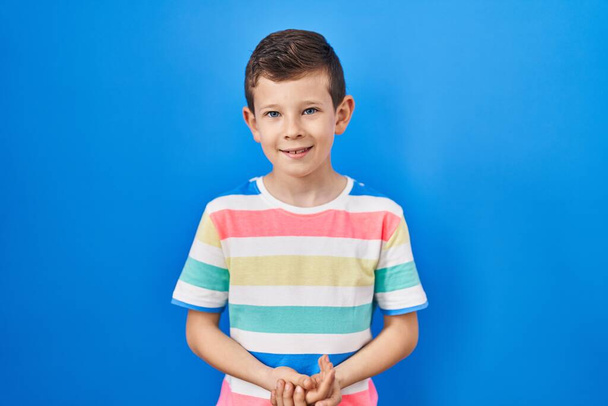 Criança caucasiana jovem em pé sobre fundo azul com as mãos juntas e dedos cruzados sorrindo relaxado e alegre. sucesso e otimista  - Foto, Imagem