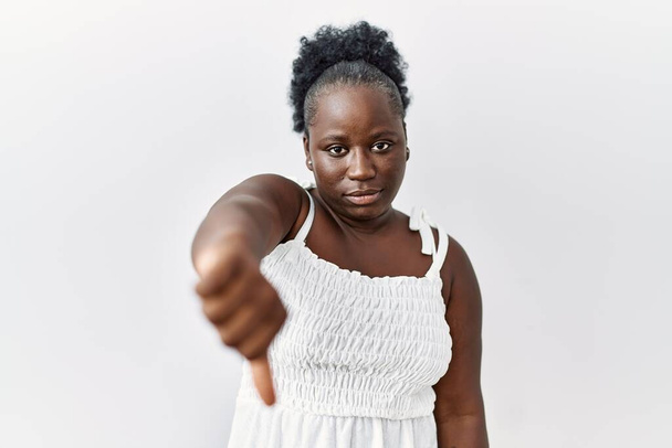 Молода африканка, що стоїть над білим ізольованим тлом, виглядає нещасною і сердитою, демонструючи відторгнення і негативні з великими пальцями вниз жестом. поганий вираз
.  - Фото, зображення