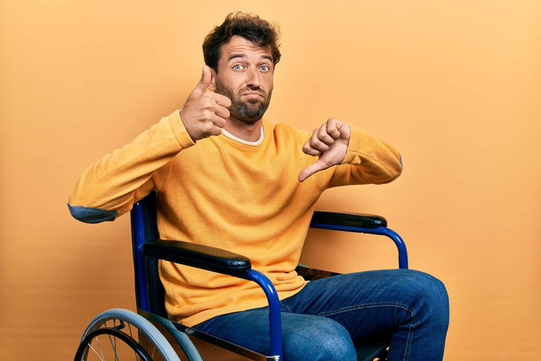 Όμορφος άντρας με γενειάδα κάθεται σε αναπηρική καρέκλα κάνει αντίχειρες πάνω και κάτω, διαφωνία και την έκφραση της συμφωνίας. τρελή σύγκρουση  - Φωτογραφία, εικόνα