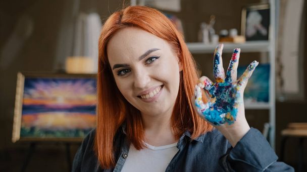 Red-haired girl talented artist female painter stands posing in art studio workshop looking at camera show okay gesture dirty palm in acrylic oil paint support great idea done successful work painting - Foto, afbeelding