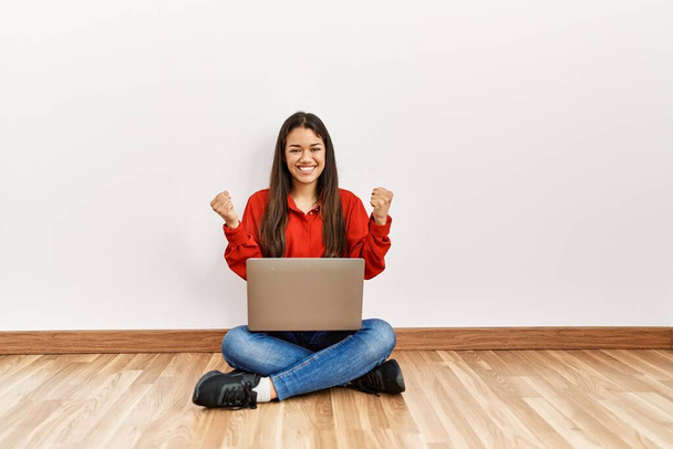 Young brunette woman sitting on the floor at empty room with laptop screaming proud, celebrating victory and success very excited with raised arms  - Photo, Image