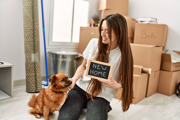 Young hispanic woman holding new home blackboard sitting on floor with dog at new home - Photo, Image