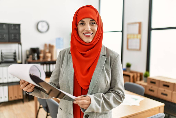 Young woman wearing arabic scarf reading document at office - Фото, изображение