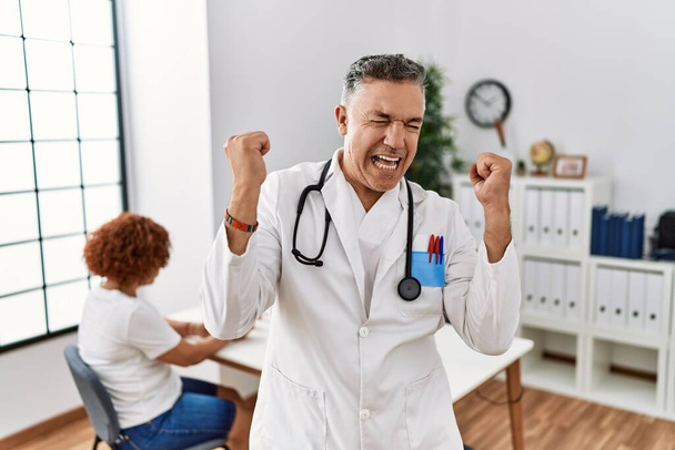 Middle age doctor man at the clinic with a patient very happy and excited doing winner gesture with arms raised, smiling and screaming for success. celebration concept.  - Photo, image