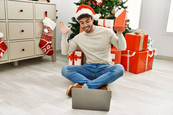 Młody Hiszpan uśmiechnięty szczęśliwy trzymając prezent świąteczny mając wideo połączenie za pomocą laptopa w domu. - Zdjęcie, obraz