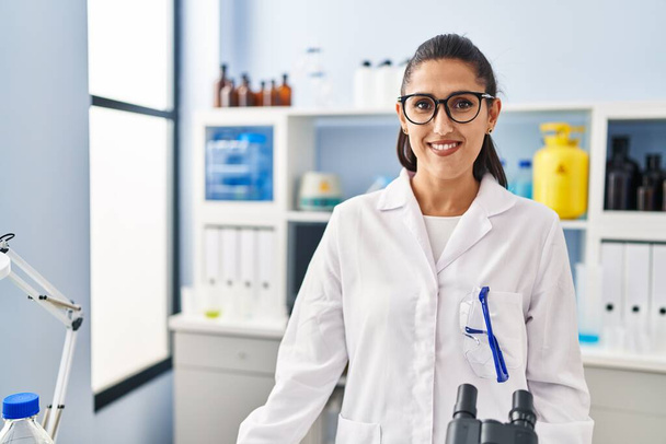 Mujer hispana joven vistiendo uniforme científico de pie en el laboratorio - Foto, Imagen