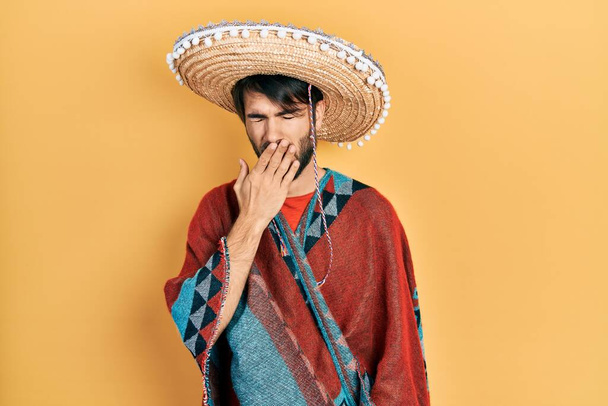 Junger hispanischer Mann mit mexikanischem Hut gelangweilt gähnend müden Mund mit der Hand bedecken. Ruhelos und schläfrig.  - Foto, Bild