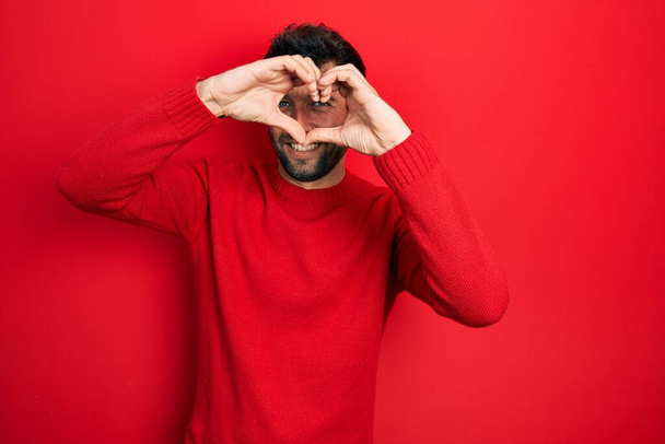Pohledný muž s vousy na sobě ležérní červený svetr dělá tvar srdce s rukou a prsty se usmívá pohled přes ceduli  - Fotografie, Obrázek