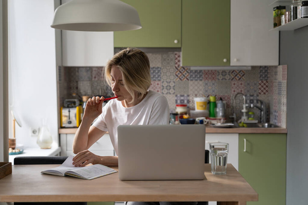 Serious middle-aged woman sits in cozy kitchen making notes in paper notebook about different job vacancies. Concentrated blonde female uses laptop for scrolling Internet recruiting websites closeup - Photo, image