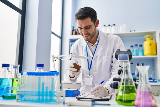 Jonge Spaanse man wetenschapper met edelsteen schrijven op document in het laboratorium - Foto, afbeelding