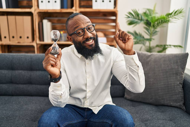 Afrikanischer Mann mit Sanduhr im Büro, lächelnd mit einer Idee oder Frage, Zeigefinger mit glücklichem Gesicht, Nummer eins  - Foto, Bild