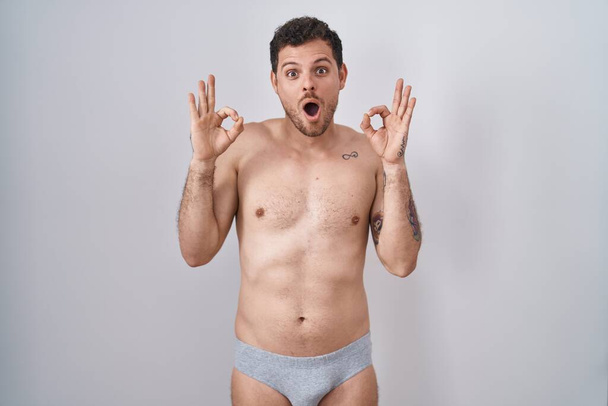 Joven hombre hispano de pie sin camisa usando ropa interior mirando sorprendido y sorprendido haciendo símbolo de aprobación ok con los dedos. expresión loca  - Foto, imagen