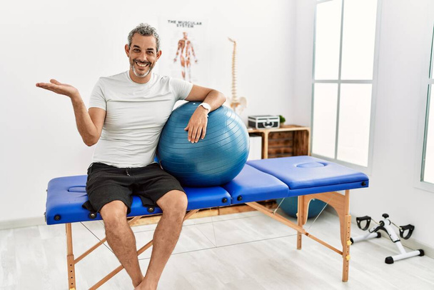Middle age hispanic man at pain recovery clinic holding pilates ball pointing aside with hands open palms showing copy space, presenting advertisement smiling excited happy  - Фото, зображення