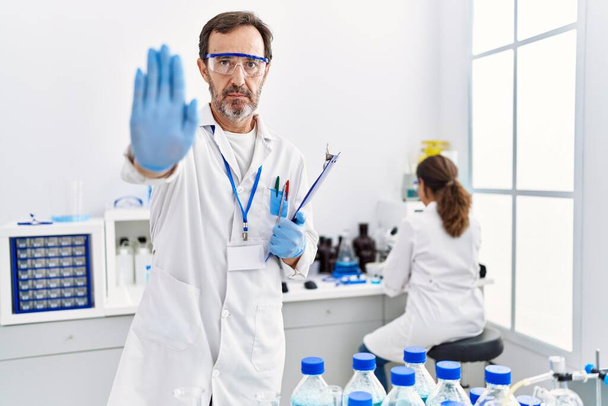 Middle age man working at scientist laboratory with open hand doing stop sign with serious and confident expression, defense gesture  - Foto, Imagem