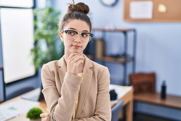 Teenager girl working at the office wearing glasses serious face thinking about question with hand on chin, thoughtful about confusing idea  - Foto, immagini