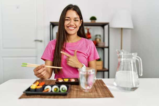 Young brunette woman eating sushi using chopsticks cheerful with a smile on face pointing with hand and finger up to the side with happy and natural expression  - Foto, immagini