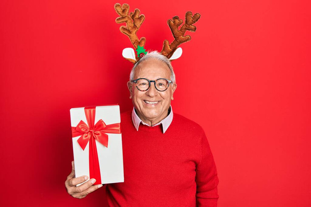 Senior man with grey hair wearing deer christmas hat holding gift looking positive and happy standing and smiling with a confident smile showing teeth  - Foto, Imagem