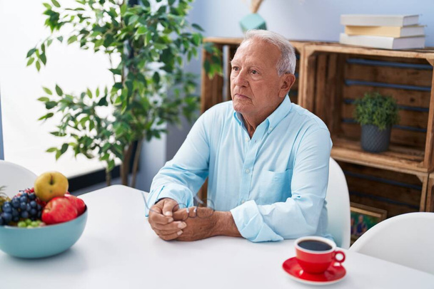 Старший седой мужчина пьет кофе сидя дома на столе - Фото, изображение