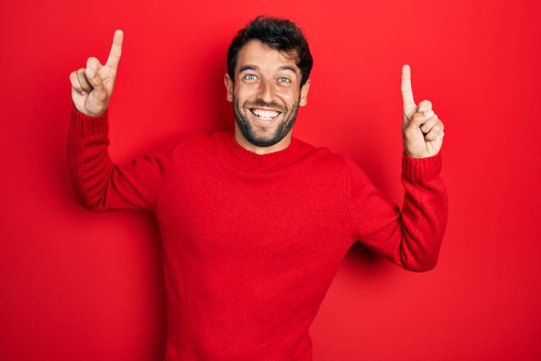 Knappe man met baard in casual rode trui glimlachend verbaasd en verrast en wijzend met vingers en opgeheven armen.  - Foto, afbeelding
