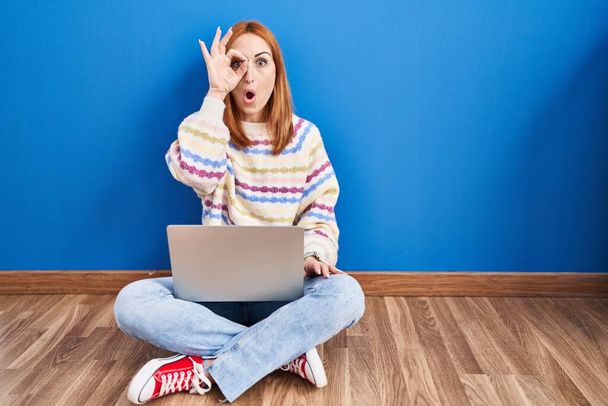Mujer joven usando el ordenador portátil en casa sentada en el suelo haciendo un gesto bien sorprendido con la cara sorprendida, los ojos mirando a través de los dedos. expresión incrédula.  - Foto, Imagen