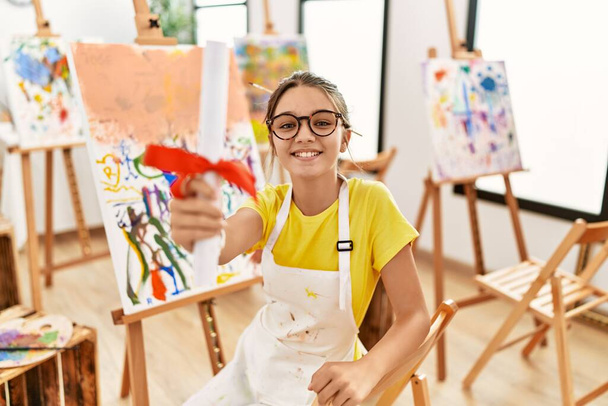 Adorável menina sorrindo confiante segurando diploma no estúdio de arte - Foto, Imagem