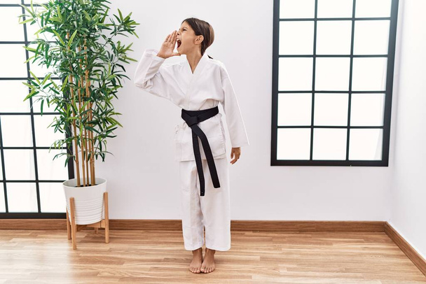 Young hispanic girl wearing karate kimono and black belt shouting and screaming loud to side with hand on mouth. communication concept.  - Photo, image