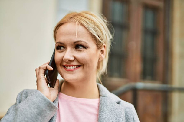 Mladý blondýnka businesswoman usměvavý šťastný mluvení na smartphone ve městě. - Fotografie, Obrázek