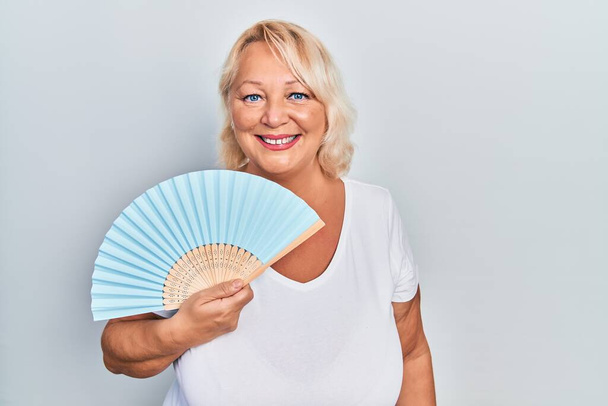 Middle age blonde woman waving hand fan cooling air in summer looking positive and happy standing and smiling with a confident smile showing teeth  - Photo, Image