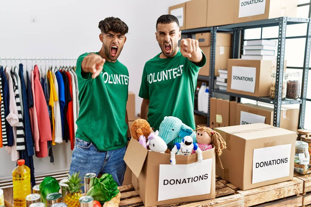 jovem gay casal vestindo voluntário t-shirt no donativos stand apontando descontente e frustrado para o câmara, irritado e furioso com você  - Foto, Imagem