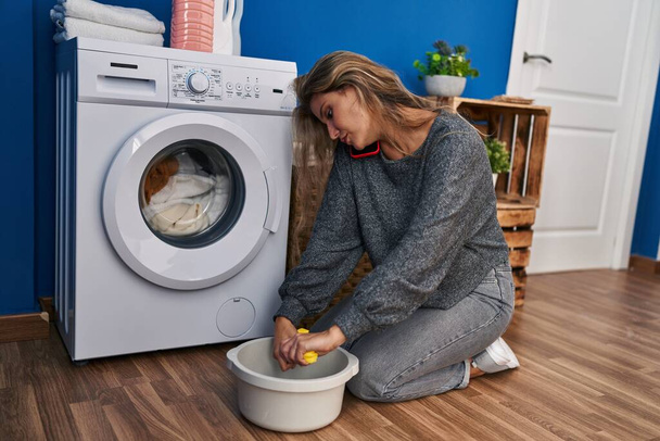 Mujer rubia joven hablando en el teléfono inteligente esperando la lavadora en la sala de lavandería - Foto, imagen