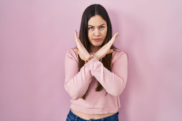 Молодая брюнетка женщина стоит на розовом фоне отвержение выражение лица скрещивание рук делает негативный знак, сердитый лицо  - Фото, изображение