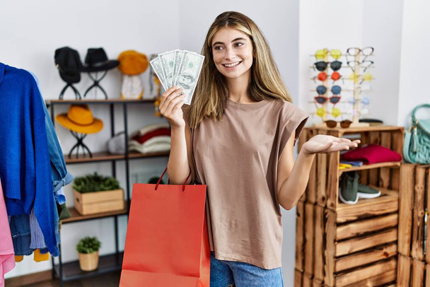 Alışveriş torbaları ve dolar tutan genç sarışın kadın başarıyı kutlamak için giyim mağazasında gülümsüyor ve kazanan yüz ifadesini kaldırıyor.  - Fotoğraf, Görsel