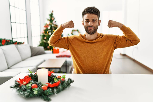 Άραβας νεαρός άνδρας κάθεται στο τραπέζι δίπλα στο χριστουγεννιάτικο δέντρο δείχνοντας τους μυς των χεριών χαμογελώντας περήφανος. έννοια της καταλληλότητας.  - Φωτογραφία, εικόνα