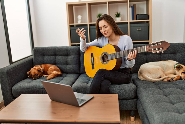 Jonge Spaanse vrouw met online gitaarles zittend op de bank met honden thuis - Foto, afbeelding