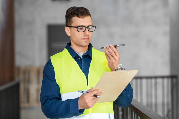 architecture, construction business and building concept - male architect in safety west with clipboard using voice command recorder on smartphone at office - Photo, Image