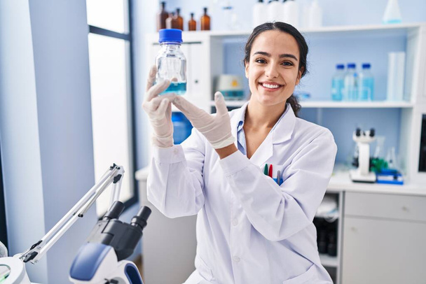 Fiatal spanyol nő, tudós egyenruhát visel, palackot tart a laborban. - Fotó, kép