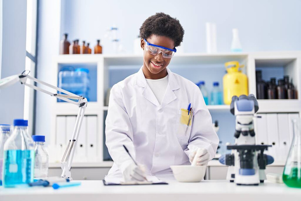 Αφροαμερικανή γυναίκα φορώντας στολή επιστήμονα γράφουν στο πρόχειρο εργασίας στο εργαστήριο - Φωτογραφία, εικόνα