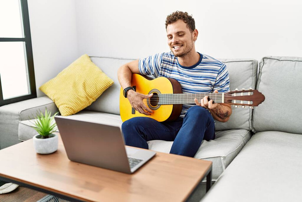 Joven hombre hispano sonriendo confiado tocando concierto de guitarra en línea en casa - Foto, imagen