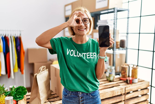 Középkorú szőke nő dolgozik visel önkéntes t ing mutatja smartphone képernyő mosolygós boldog csinál oké jel kézzel a szemen keresztül néz az ujjak  - Fotó, kép