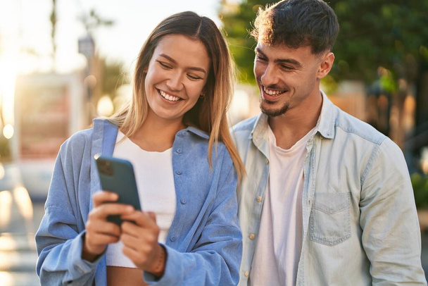 Молодой мужчина и женщина улыбаются уверенно, используя смартфон на улице - Фото, изображение