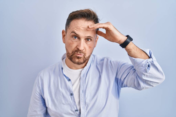 Middle age caucasian man standing over blue background worried and stressed about a problem with hand on forehead, nervous and anxious for crisis  - Foto, imagen