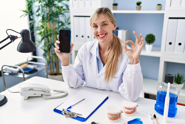 Beautiful dentist woman showing smartphone screen doing ok sign with fingers, smiling friendly gesturing excellent symbol  - Photo, image