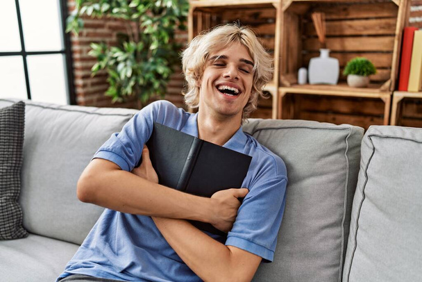 Young blond man hugging book sitting on sofa at home - 写真・画像