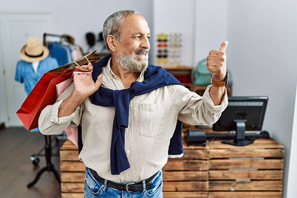 Handsome senior man holding shopping bags at boutique shop smiling with happy face looking and pointing to the side with thumb up.  - Foto, imagen