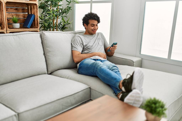 Νεαρός ισπανόφωνος άνδρας που χρησιμοποιεί smartphone κάθεται στον καναπέ στο σπίτι. - Φωτογραφία, εικόνα