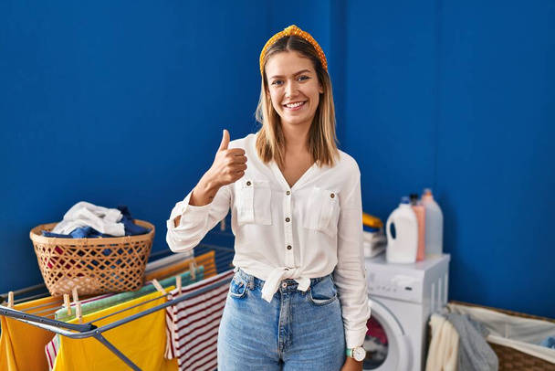 Young blonde woman at laundry room doing happy thumbs up gesture with hand. approving expression looking at the camera showing success.  - Fotografie, Obrázek