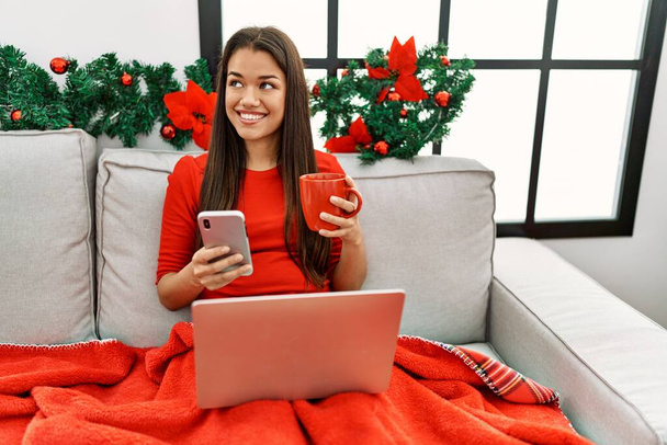 Νεαρή Λατίνα γυναίκα που χρησιμοποιεί το smartphone κάθεται από τη διακόσμηση των Χριστουγέννων στο σπίτι - Φωτογραφία, εικόνα