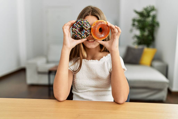 Adorable girl holding donuts over eyes sitting on table at home - Foto, Imagen