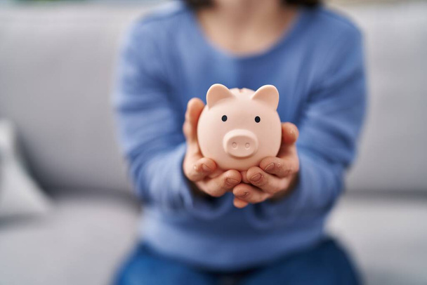 Young woman with down syndrome holding piggy bank sitting on sofa at home - Foto, Imagem