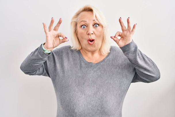Μέση ηλικία καυκάσιος γυναίκα στέκεται πάνω από το λευκό φόντο αναζητούν έκπληκτος και σοκαρισμένος κάνει εντάξει σύμβολο έγκρισης με τα δάχτυλα. τρελή έκφραση  - Φωτογραφία, εικόνα